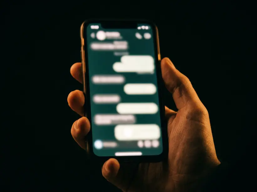Main tenant un téléphone dans le noir, écran allumé sur une conversation de messagerie